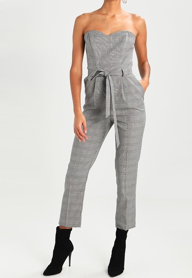 Jumpsuit w kratę w odcieniu szarym bez ramiączek, z przewiązanym paskiem w talii, z krótkimi spodniami i bocznymi kieszeniami, noszony z czarnymi botkami na wysokim obcasie.