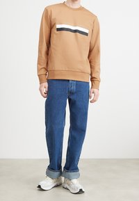 Felpa beige con logo testurizzato e tre righe orizzontali in nero, bianco e grigio. Abbinata a jeans blu larghi e sneaker bianche.