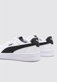 Zapatillas blancas con acentos laterales negros, que presentan un upper suave similar al cuero, puntera redonda y suela de goma texturizada. Logo de Puma en el talón.