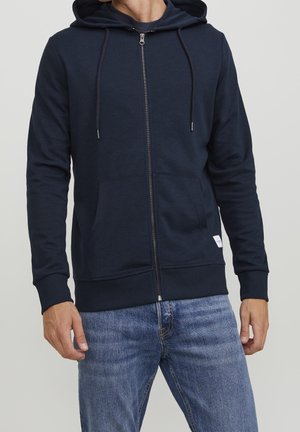 Mann trägt dunkelblauen Kapuzenpullover mit Reißverschluss und Vorderentaschen sowie hellblaue Jeans vor einfachem Hintergrund.