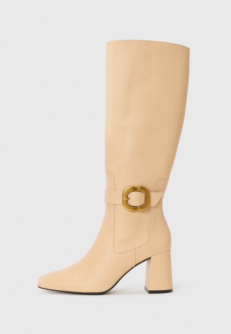 Botte en cuir beige, arrivant au genou, avec un bout carré, un talon épais, et une large ceinture avec une boucle circulaire en ton doré. Texture lisse et coutures minimales.
