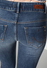 Jean en denim bleu avec une coupe ajustée, présentant des coutures contrastées, deux poches arrière et une petite étiquette en cuir sur la taille.