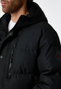 Veste matelassée noire avec capuche, dotée d'une poche zippée et d'un logo brodé sur la manche. Le matériel a une finition lisse et mate.
