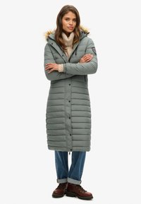 Abrigo de invierno acolchado gris con capucha forrada de pelo, con cremallera frontal y botones, combinado con vaqueros azules y botas marrones.