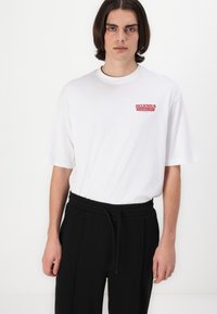 White cotton t-shirt with short sleeves and a red printed text "DELICIOUS" on the chest. Worn with black sweatpants and a drawstring waist.