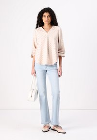 Blusa lunga beige chiara a maniche lunghe con motivo a pois dorati, abbinata a jeans a zampa d'elefante blu chiaro. Accessoriata con una borsa bianca e scarpe beige.