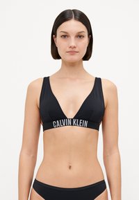 Fekete bikini felső mély V-nyakkivágással, széles elasztikus pánttal, rajta "CALVIN KLEIN" felirattal fehér színben. Simás textúra, minimalista stílus.