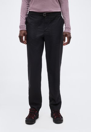 Személy fekete egyenes szárú nadrágot övhurkokkal és fekete túracipőt piros fűzőkkel visel, fehér háttér előtt áll.