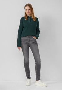 Pull en tricot côtelé vert, jeans slim gris foncé, et baskets blanches à plateforme. Le mannequin se tient avec les mains dans les poches, souriant devant un fond neutre.