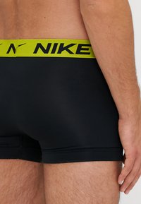 Czarne bokserki o gładkiej fakturze, z żółtym logo Nike na pasku. Krój jest dopasowany, sięgający do połowy uda.