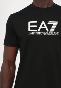 Sort bomulds T-shirt med korte ærmer, der har hvid broderet tekst 'EA7 EMPORIO ARMANI' på forsiden og horisontale stribedetaljer.