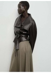 Veste en cuir marron avec un col oversize, taille ceinturée et texture lisse, associée à une jupe beige à carreaux.