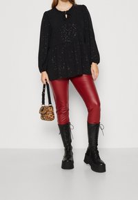 Blouse noire scintillante à manches longues avec un détail de nœud en trou de serrure, associée à des leggings en cuir synthétique rouges et des bottes de combat noires à lacets.