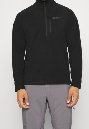 Mann trägt schwarzen Columbia Pullover-Fleece mit Reißverschluss-Brusttasche und graue Hose, steht vor neutralem Hintergrund.