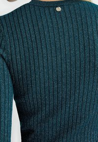 Dunkelblauer, gerippter Pullover mit metallischen Fäden, runder Ausschnitt und einem kleinen Knopfdetail auf der Rückseite. Weiche Textur mit vertikalen Streifen.