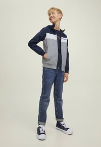 Pilko ir tamsiai mėlynos spalvos striukė su užtrauktuku ir balta juosta, dėvima virš tamsios palaidinės, derinama su mėlynais džinsais ir tamsiai mėlynais sportiniais batais. Lygus audinys, laisvas stilius.