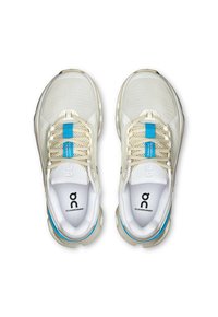 Cloudrunner-skor i ljus beige med ett mesh-överkinn, blå accenter på tungan och framträdande dämpning i sulan. Logotyp på hälen och innersulan.