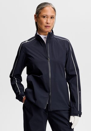 Frau trägt eine marineblaue Reißverschlussjacke mit weißen Paspeln und passende Hose, steht mit einer Hand in der Tasche und weißem Handschuh an der anderen Hand.