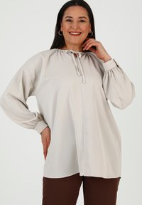 Blusa beige chiaro con scollatura rotonda e dettaglio con fiocco, caratterizzata da maniche lunghe voluminose con polsini arricciati. Tessuto morbido e vestibilità comoda.
