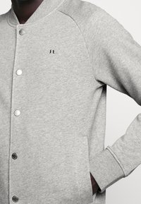 Gray sweatshirt with a baseball jacket style, featuring silver snap buttons, ribbed cuffs, side pockets, and a small logo on the chest.