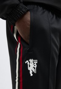 Käsi mustade trennipükste taskus, mille külgedel on valged, punased ja mustad triibud ning tasku lähedal on tikitud valge kurat tridenti hoidmas.