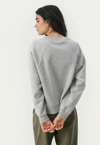 Grau Sweatshirt mit Raglan-Design, gerippten Bündchen und Saum. Getragen mit olivgrünen Hosen, von hinten gesehen, Hände hinter dem Rücken verschränkt.