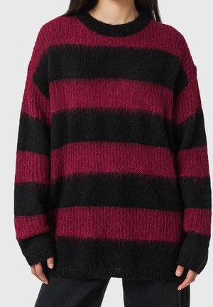Femme portant un pull ample en maille avec de larges rayures horizontales en rouge foncé et noir, associé à un pantalon sombre.