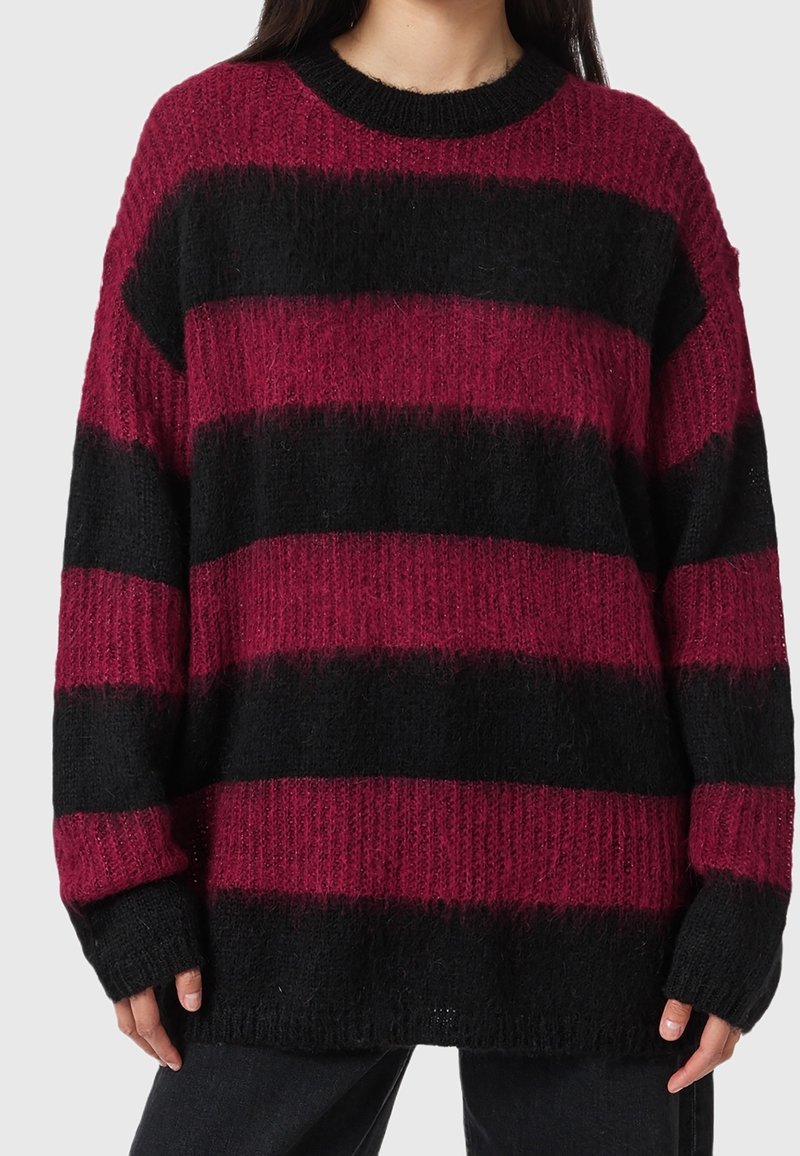 Femme portant un pull ample en maille avec de larges rayures horizontales en rouge foncé et noir, associé à un pantalon sombre.