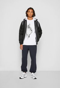 Jachetă neagră tip windbreaker, tricou alb cu grafică ce prezintă o amprentă de mână, pantaloni de trening bleumarin și adidași albi cu accente argintii.