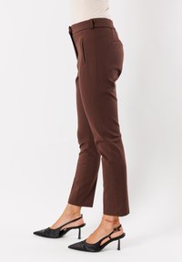 Pantaloni brown slim-fit indossati con décolleté nere a punta su una persona in piedi contro uno sfondo bianco.