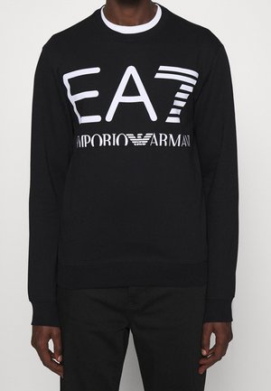 Mann trägt ein schwarzes Langarmshirt mit weißem EA7 Emporio Armani-Logo auf der Brust und weißem Kragenabschluss.