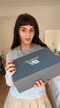 Jovem mulher com cabelo escuro segura uma caixa de sapatos da New Balance, de pé em um quarto com paredes claras e piso de madeira.