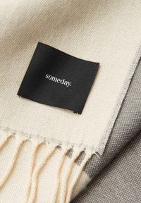 Sjaal van wolmix in crème- en grijstinten, met rafelige randen. Zwart label met witte tekst waarop "someday" staat.