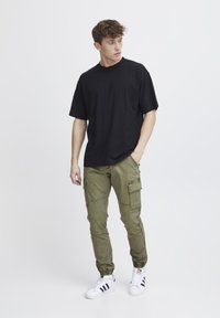 T-shirt noir à manches courtes, pantalon cargo olive avec poches, chaussures blanches avec rayures noires, se tenant devant un fond clair.
