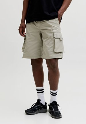 Mann trägt beige Cargo-Shorts, schwarze Sneaker, weiße Socken mit schwarzen Streifen und ein schwarzes Shirt, steht mit den Händen in den Taschen.
