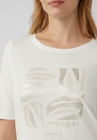 T-shirt blanc à manches courtes avec des motifs géométriques dorés métalliques et le texte "Accepte ton unicité" imprimé en bas.