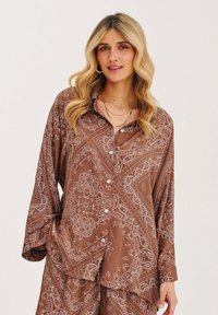 Hnědá košile na knoflíky s bílými vzory paisley, dlouhé rukávy a uvolněný střih. Tkanina vypadá hladce a lehce.