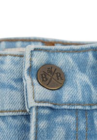 Ljust blå denimtyg med en synlig metallknapp som har korsade symboler och präglade bokstäver "B/R", samt kontrasterande sömnad.