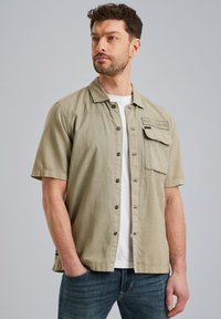 Kortærmet skjorte i oliven grøn med knaplukning, brystlomme og broderet labeldetalje, parret med en hvid t-shirt og blå jeans.