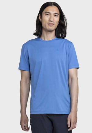 Junger Mann mit langen schwarzen Haaren, der ein hellblaues kurzärmeliges Hemd und dunkle Hosen trägt, vor einem schlichten hellen Hintergrund stehend.