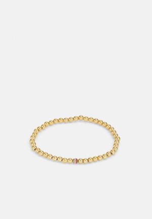 Złota, rozciągliwa bransoletka z koralików z jednym pasiastym koralikiem w czerwone, białe i czarne akcenty na jednolitym jasnoszarym tle.