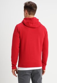 Sudadera con capucha roja de textura suave, con puños y dobladillo acanalados, que cuenta con un bolsillo tipo canguro. Diseño liso, sin logotipos ni patrones visibles.