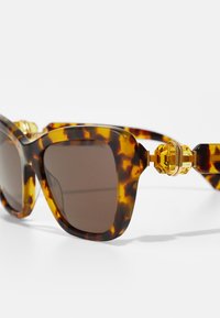 Ochelari de soare din țestoasă cu lentile maronii, rame voluminoase și accente din aur, având un design geometric pe brațele ochelarilor.