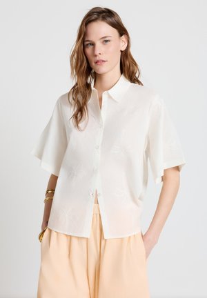 Jeune femme aux cheveux bruns détachés, portant une chemise blanche à manches courtes boutonnée et un pantalon taille haute couleur pêche clair, posant avec les mains dans les poches.