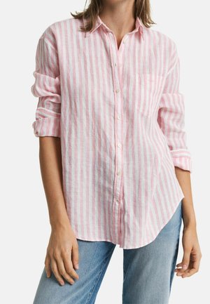 Femme portant une chemise boutonnée à manches longues à rayures verticales roses et blanches, manches retroussées, et un jean bleu clair.
