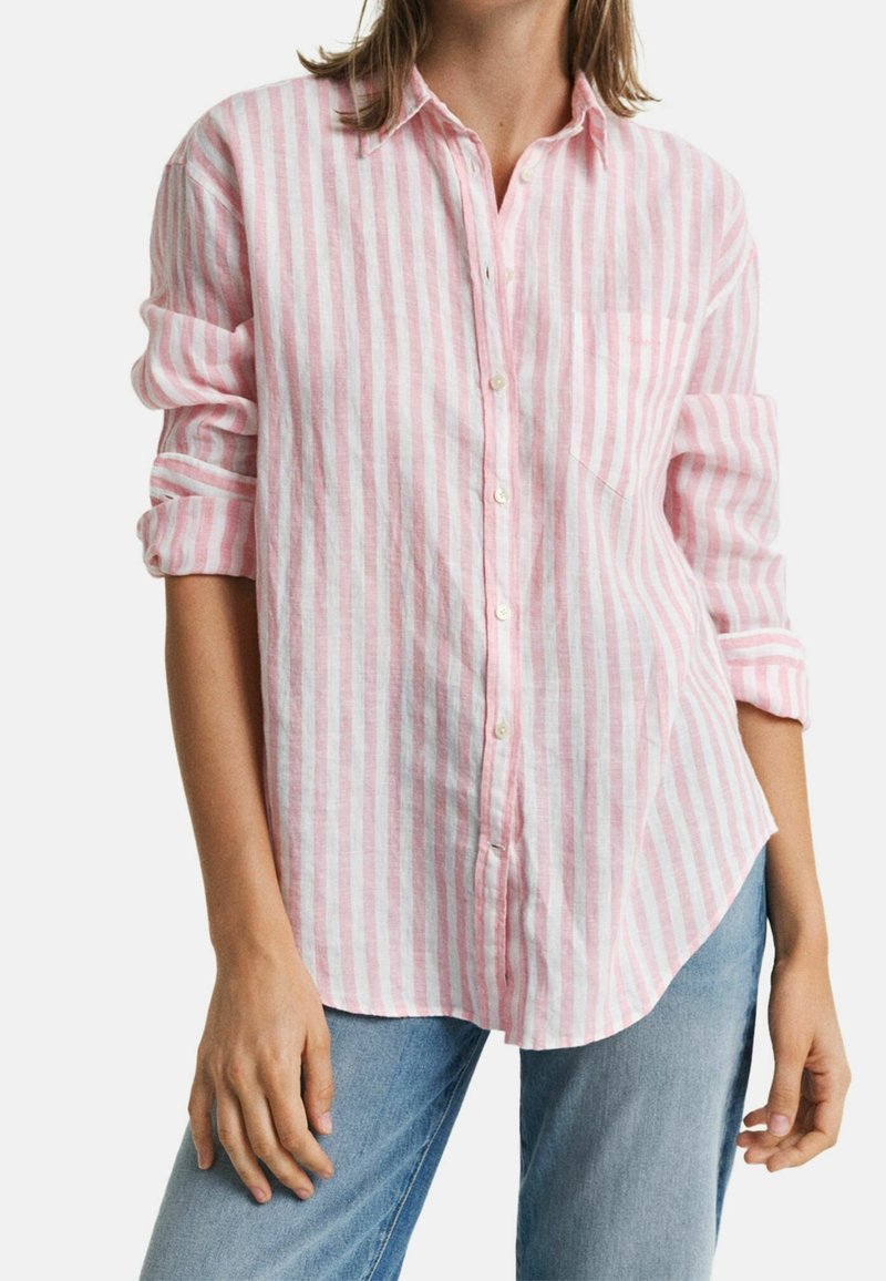 Femme portant une chemise boutonnée à manches longues à rayures verticales roses et blanches, manches retroussées, et un jean bleu clair.