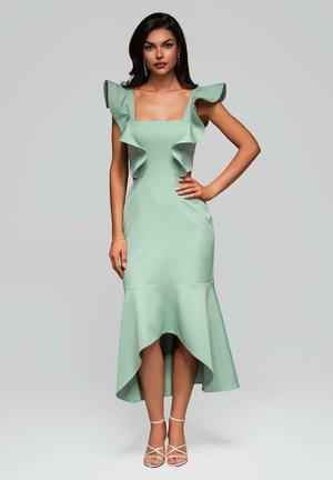 Mint green fitted dress with ruffled shoulder accents and a high-low hem. Smooth, textured fabric. Paired with strappy light-colored heels.