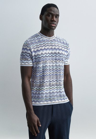 Homme portant une chemise à manches courtes avec un motif en zigzag multicolore et un pantalon foncé texturé, debout devant un fond clair uni.