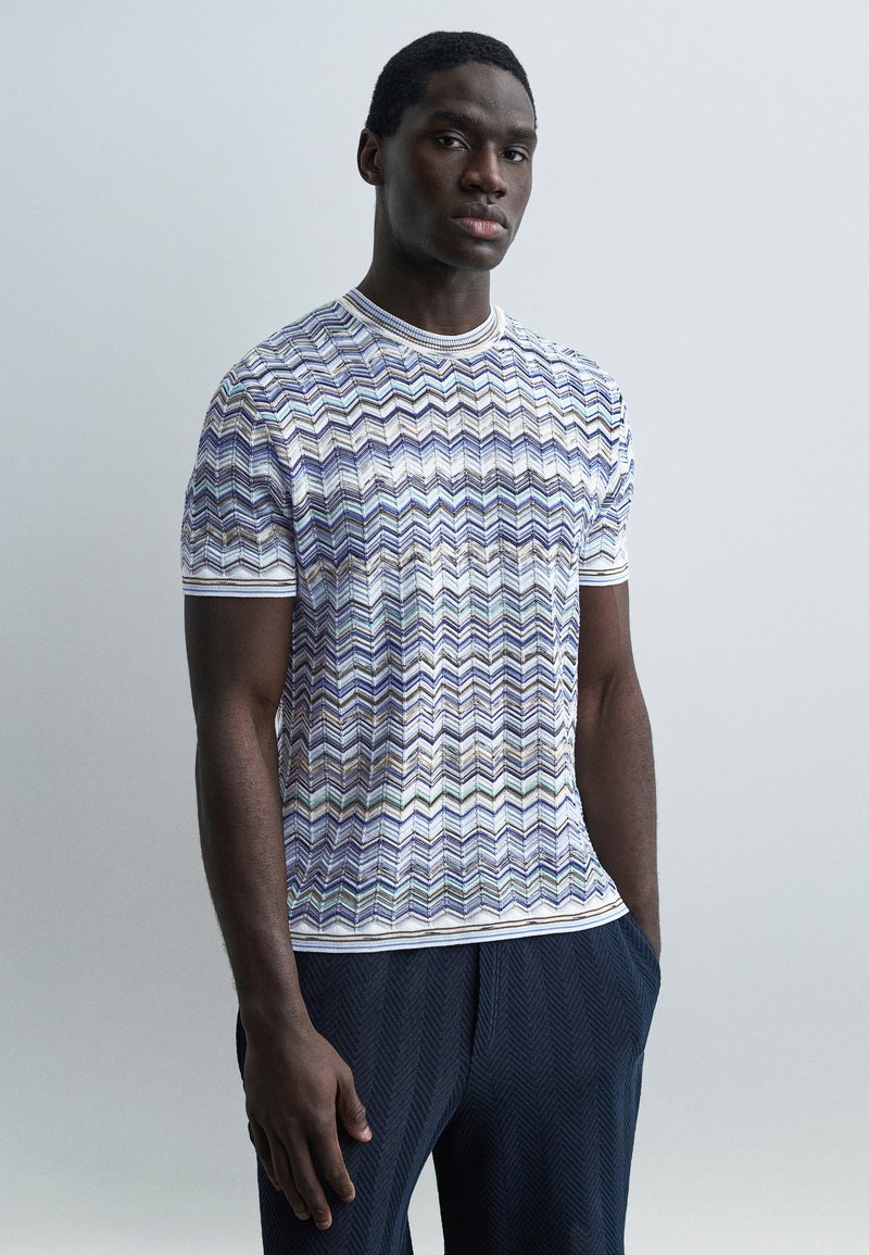 Άντρας που φοράει κοντομάνικο πουκάμισο με πολύχρωμο ζιγκ-ζαγκ σχέδιο και σκούρο παντελόνι με υφή, στέκεται μπροστά σε απλό φωτεινό φόντο.