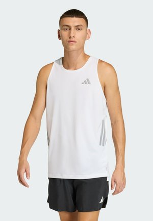 Jeune homme portant un débardeur blanc de sport et un short noir avec des logos Adidas, debout devant un fond clair uni.
